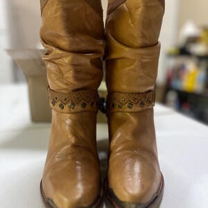 May include: A pair of tan mid-calf boots featuring a slouchy shaft, a textured upper cuff, and a decorative strap across the ankle adorned with small metal studs and diamond-shaped accents. The boots have a pointed toe with a metallic toe cap.