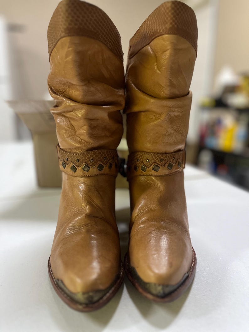 May include: A pair of tan mid-calf boots featuring a slouchy shaft, a textured upper cuff, and a decorative strap across the ankle adorned with small metal studs and diamond-shaped accents. The boots have a pointed toe with a metallic toe cap.