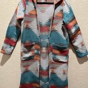 May include: A long, open-front cardigan with a Southwestern-inspired pattern in shades of gray, teal, red, and orange. The hooded sweater features two front pockets and the brand name "DICTIONARY" on the label.