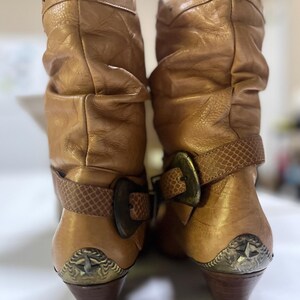 May include: A pair of tan slouchy mid-calf boots featuring a textured strap with a large bronze-toned buckle across the ankle. The boots have a row of small studs along the top edge and a metallic heel cap detail with a star emblem on the back.