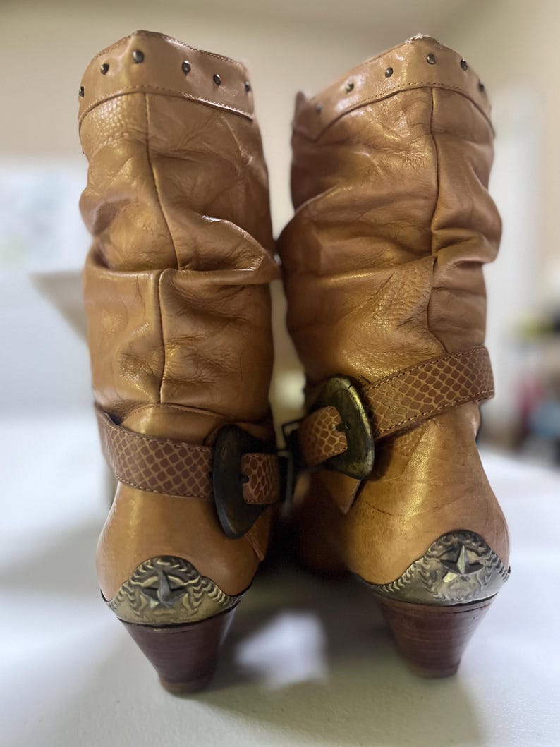 May include: A pair of tan slouchy mid-calf boots featuring a textured strap with a large bronze-toned buckle across the ankle. The boots have a row of small studs along the top edge and a metallic heel cap detail with a star emblem on the back.
