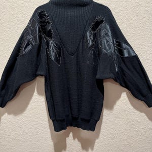 May include: Black sweater with a high collar and decorative leaf-shaped embellishments on the shoulders. The sweater has long sleeves and a ribbed texture on the collar and lower hem.