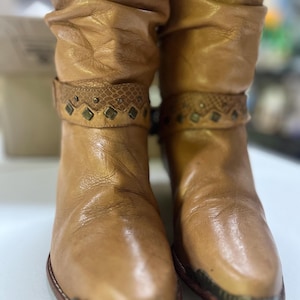 May include: A pair of tan leather slouch boots featuring a pointed toe with a metallic cap. Each boot has a decorative strap around the ankle with a snakeskin-textured band, small metal studs, and diamond-shaped accents in a dark finish.