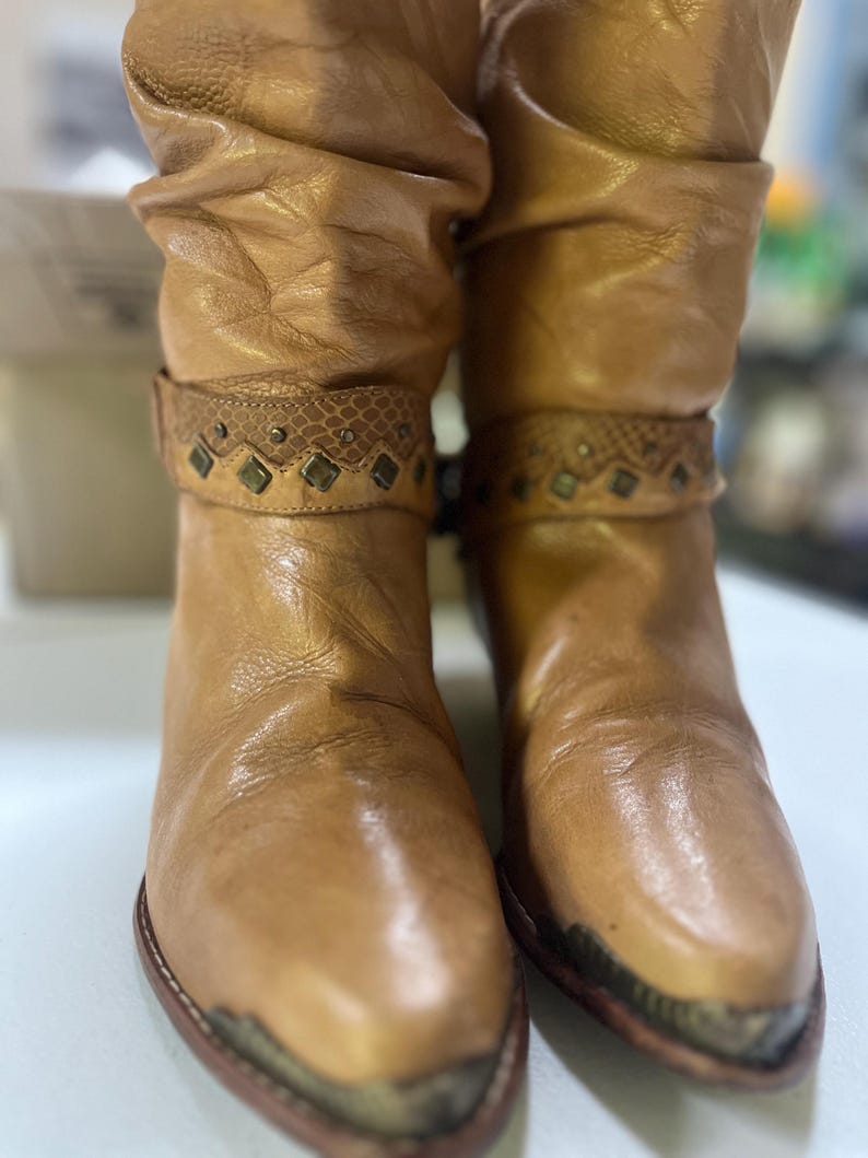 May include: A pair of tan leather slouch boots featuring a pointed toe with a metallic cap. Each boot has a decorative strap around the ankle with a snakeskin-textured band, small metal studs, and diamond-shaped accents in a dark finish.
