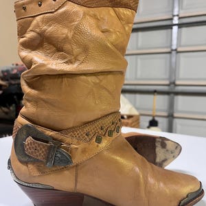 May include: A tan leather mid-calf boot featuring a slouchy shaft, a textured snakeskin-patterned collar, and a decorative strap with a large antiqued metal buckle. The boot includes small metal studs, a pointed toe with a metal cap, and a wooden heel.