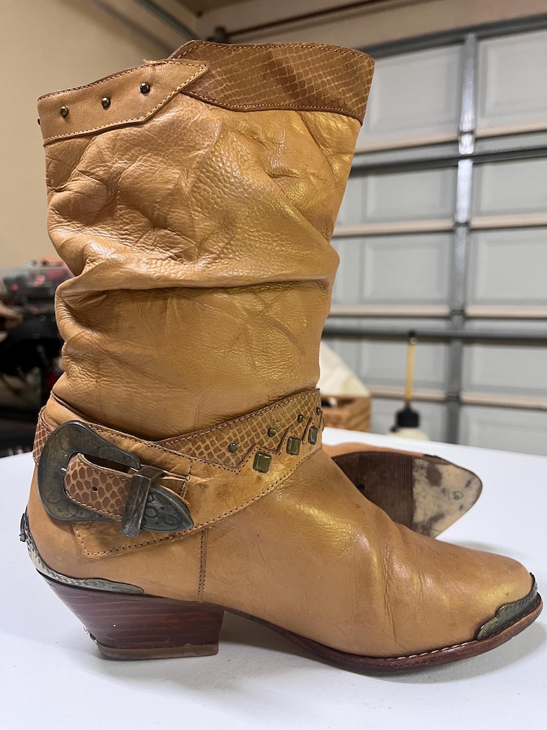 May include: A tan leather mid-calf boot featuring a slouchy shaft, a textured snakeskin-patterned collar, and a decorative strap with a large antiqued metal buckle. The boot includes small metal studs, a pointed toe with a metal cap, and a wooden heel.