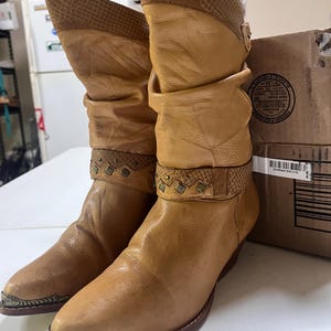 May include: A pair of tan leather mid-calf boots featuring a slouchy shaft, a textured snakeskin-patterned collar, and a decorative strap across the ankle with small metal studs. The boots have a pointed toe with a metallic silver-toned toe cap.