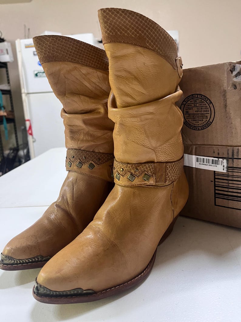 May include: A pair of tan leather mid-calf boots featuring a slouchy shaft, a textured snakeskin-patterned collar, and a decorative strap across the ankle with small metal studs. The boots have a pointed toe with a metallic silver-toned toe cap.