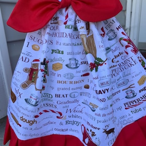 May include: A white hanging kitchen towel featuring a red fabric bow at the top and a red ruffled hem at the bottom. The towel is printed with a holiday-themed recipe pattern including gingerbread men, candy canes, and text instructions.