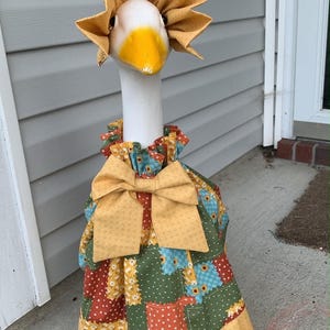 May include: A white decorative goose statue wearing a yellow bonnet and a patchwork dress featuring green, red, and blue floral patterns. The dress has a ruffled collar, a large yellow bow at the neck, and a pleated yellow hem at the bottom.