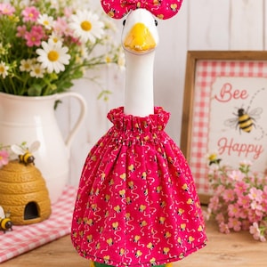 May include: A ceramic goose figurine wearing a pink dress and bow, both adorned with a bee pattern. The goose has a yellow beak and feet, and the dress has a ruffled collar. The background includes a framed sign that says "Bee Happy".