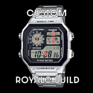 Könnte beinhalten: Eine silberfarbene Digitalarmbanduhr mit schwarzem Zifferblatt und der Aufschrift CUSTOM ROYALE BUILD. Das Display zeigt ein kreisförmiges analoges Hilfszifferblatt, eine Weltkarte und die Uhrzeit 10:48 sowie die Beschriftungen 5 ALARMS und 10 YEAR BATTERY.