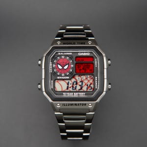 Puede incluir: Reloj digital Casio plateado con diseño de Spider-Man en la esfera. El reloj tiene una pantalla digital negra y roja que muestra la hora. La correa del reloj es de metal plateado.