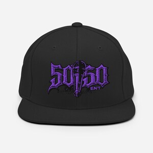 Puede incluir: Gorra de béisbol negra con visera plana. La gorra presenta el texto morado "50/50 ENT" en una fuente estilizada. El diseño incluye un gráfico central que se asemeja a una antorcha o micrófono.