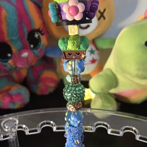 以下が含まれることがあります： 装飾的なビーズデザインの青いペン。ペンは、緑と茶色のビーズを含む一連のカラフルなビーズと、上部にレゴのようなフィギュアが特徴です。ペンは、さまざまなチャームと花で装飾されています。