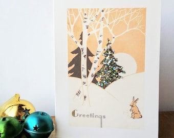 May include: A vintage Christmas card with a snowy scene featuring two birch trees, a glitter-covered evergreen tree, and a small rabbit. The card has the word "Greetings" printed in a decorative font.