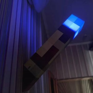 Könnte beinhalten: Eine Minecraft-Themen-Wandlampe mit pixeligem Design. Die Lampe ist rechteckig mit einem blau leuchtenden Oberteil und einem holzähnlichen Körper. Sie ist an einer Wand mit gestreifter Tapete befestigt und bildet ein einzigartiges Dekorationselement.