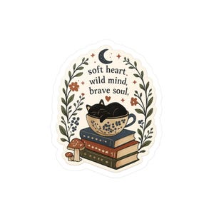 Może przedstawiać: Naklejka przedstawiająca czarnego kota śpiącego w kwiatowej filiżance na stosie książek. Naklejka zawiera słowa "soft heart, wild mind, brave soul" i jest otoczona kwiatową obwódką z grzybem na dole.