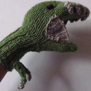 Op de afbeelding: Groene gebreide dinosauruspop met een grijze mond en een zwart oog. De pop wordt op de hand van een persoon gedragen.