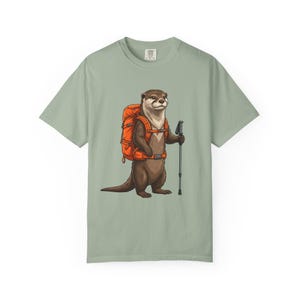 Op de afbeelding: Een saliegroen t-shirt met een grafische illustratie van een bruine otter die rechtop staat. De otter draagt een feloranje wandelrugzak en houdt een zwarte wandelstok in zijn rechterpoot, afgebeeld in een gedetailleerde cartoonstijl.