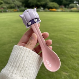 May include: A pink ceramic spoon with a cartoon character head at the top. The character has a purple head, purple eyes, and a purple tongue. The spoon is held in a hand with a white sweater sleeve in the background.