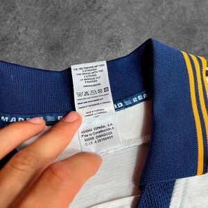 May include: Close-up of a white and navy blue athletic jersey collar with yellow stripes. The jersey tag displays text in multiple languages, including "THE BRAND WITH THE 3 STRIPES" and "100% POLYESTER".