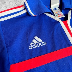 May include: A blue Adidas sports shirt with white stripes and a red stripe across the chest. The Adidas logo is embroidered in white. The shirt has a collar and a tag with the Adidas logo.