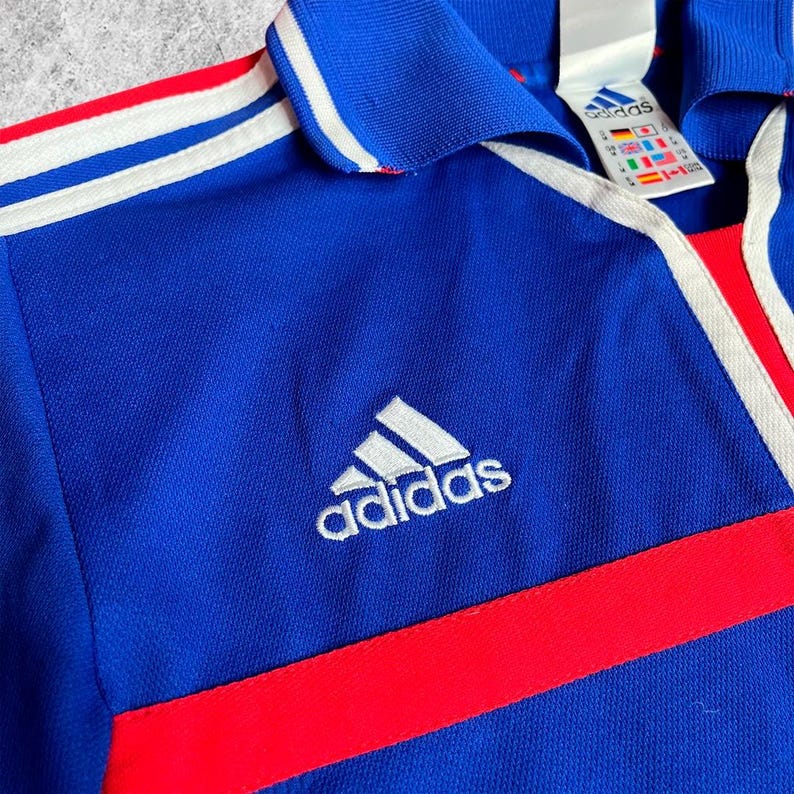 May include: A blue Adidas sports shirt with white stripes and a red stripe across the chest. The Adidas logo is embroidered in white. The shirt has a collar and a tag with the Adidas logo.