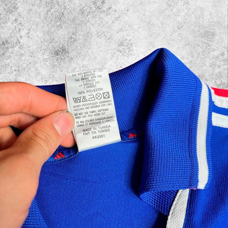 May include: A close-up of a blue athletic shirt with white stripes and a red accent. A tag with text in multiple languages is held up, showing care instructions and the brand's logo. The shirt is made of 100% polyester.