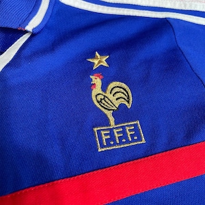 May include: Close-up of a blue jersey featuring a gold rooster emblem, a gold star, and the letters "F.F.F." in a gold rectangle. The jersey has a red stripe and white stripes on the collar.