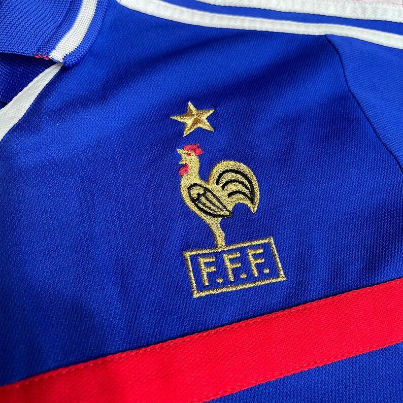 May include: Close-up of a blue jersey featuring a gold rooster emblem, a gold star, and the letters "F.F.F." in a gold rectangle. The jersey has a red stripe and white stripes on the collar.