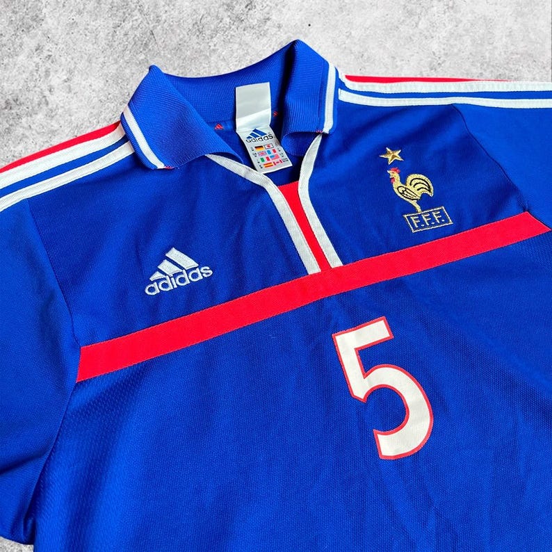 May include: Blue football shirt with red and white detailing. Includes the Adidas logo, a French team emblem, and the number 5 in white. The shirt has a collared neckline and white stripes on the sleeves.