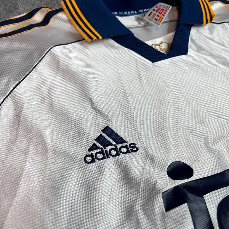 May include: White soccer jersey with navy blue and gold accents. Features the Adidas logo in navy blue on the chest. The collar and sleeve stripes are navy blue with gold trim. The jersey has a V-neck design.