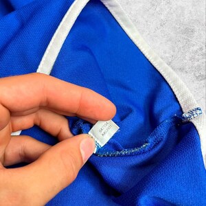 May include: Close-up of a blue athletic shirt with white trim. The shirt's tag is visible, displaying the numbers "647194" and "AKU001". The fabric has a textured appearance.