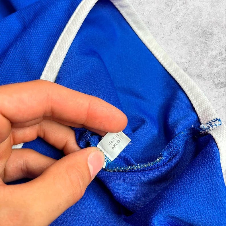 May include: Close-up of a blue athletic shirt with white trim. The shirt's tag is visible, displaying the numbers "647194" and "AKU001". The fabric has a textured appearance.