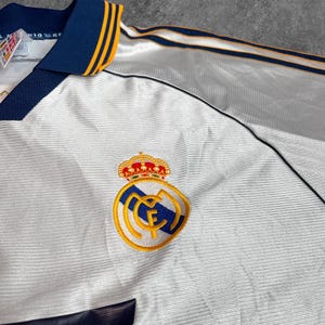 May include: White soccer jersey with navy blue and gold accents. Features a team crest with a crown and the team's logo. The collar and sleeves have navy blue and gold trim.