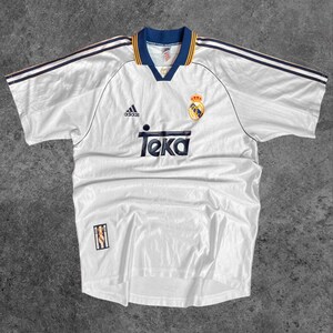 May include: White soccer jersey with navy blue and yellow accents. Features the Adidas logo, the Teka sponsor logo, and a team crest. The jersey has a V-neck collar and navy blue stripes on the shoulders.