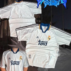 May include: White soccer jersey with blue and yellow trim, featuring the Adidas logo and "Teka" text. The jersey has short sleeves and a V-neck collar. A crest is visible on the chest. The jersey is likely vintage.