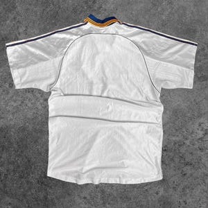 May include: A white athletic jersey with a blue and yellow collar and blue stripes on the shoulders. The back of the jersey is visible, showing the curved yoke and the seams. The fabric has a slight sheen.