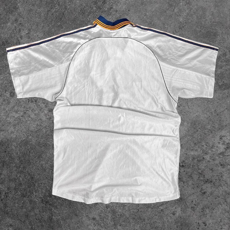 May include: A white athletic jersey with a blue and yellow collar and blue stripes on the shoulders. The back of the jersey is visible, showing the curved yoke and the seams. The fabric has a slight sheen.