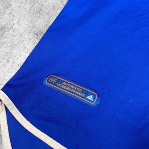 May include: A close-up of a blue athletic garment with white trim. The garment has a patch that reads "FFF AUTHENTIC LICENSED PRODUCT" with the Adidas logo. The fabric appears to be a smooth, athletic material.