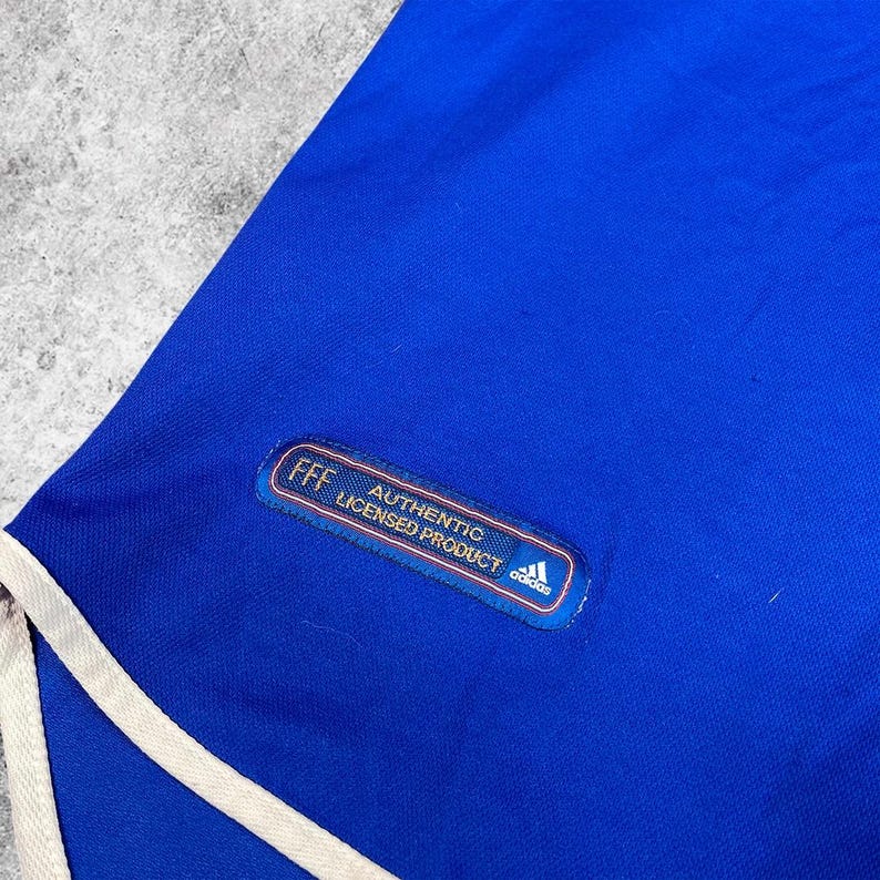 May include: A close-up of a blue athletic garment with white trim. The garment has a patch that reads "FFF AUTHENTIC LICENSED PRODUCT" with the Adidas logo. The fabric appears to be a smooth, athletic material.