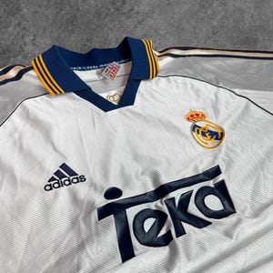 May include: White vintage soccer jersey with navy blue and gold accents. Features the Adidas logo, the Teka sponsor logo, and a team crest. The collar and sleeves have navy blue and gold trim.