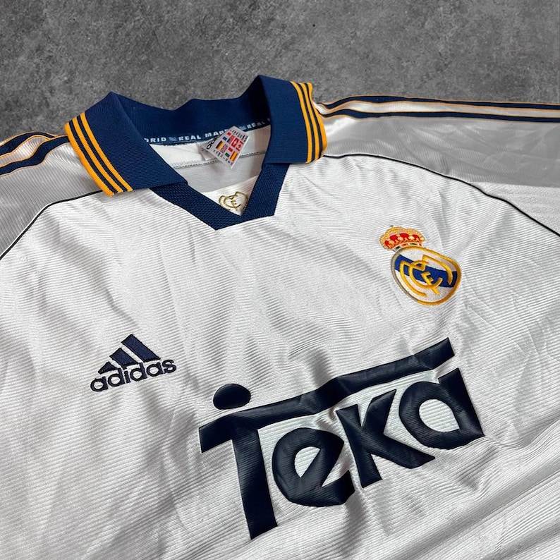 May include: White vintage soccer jersey with navy blue and gold accents. Features the Adidas logo, the Teka sponsor logo, and a team crest. The collar and sleeves have navy blue and gold trim.