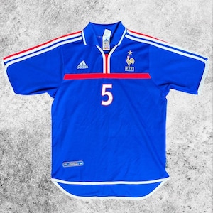 May include: A blue football shirt with white and red detailing. The shirt displays the number 5 in white, the Adidas logo, and the French Football Federation emblem. It has a collar and short sleeves.