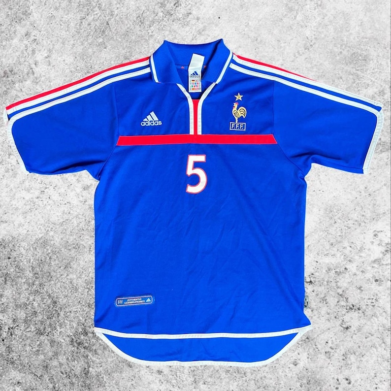 May include: A blue football shirt with white and red detailing. The shirt displays the number 5 in white, the Adidas logo, and the French Football Federation emblem. It has a collar and short sleeves.