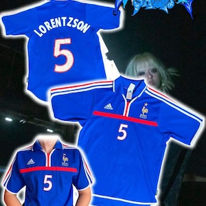 May include: Three blue football shirts with white and red detailing. One shirt has the name "LORENTZSON" and the number "5" on the back. The other two shirts have the number "5" on the front. The Adidas logo is visible on two of the shirts.