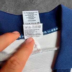 May include: Close-up of a white and blue garment tag with text in multiple languages. The tag includes washing instructions and the phrase "DIE WELTMARKE MIT DEN 3 STREIFEN". The garment is made of 100% polyester.