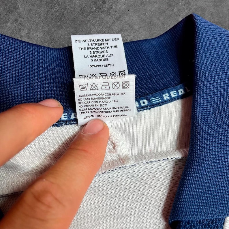 May include: Close-up of a white and blue garment tag with text in multiple languages. The tag includes washing instructions and the phrase "DIE WELTMARKE MIT DEN 3 STREIFEN". The garment is made of 100% polyester.