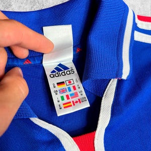 May include: A close-up of a blue Adidas jersey with white stripes and a red accent. The jersey features a white tag with the Adidas logo and country flags. The collar is blue with a white drawstring.